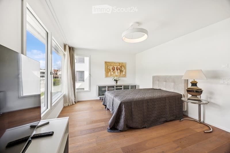 Modern bedroom with wooden flooring, large windows, a bed with a grey duvet, a nightstand with a lamp, a small table, and a painting on the wall.