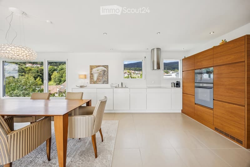 Modern kitchen with white cabinets, a wooden dining table, four striped chairs, and a view of greenery through large windows.