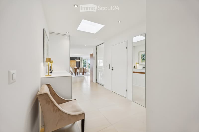 Bright, modern hallway with white walls, recessed lighting, and a skylight. Features a small seating area with a chair and a console table with lamps, leading to a dining area visible in the background.