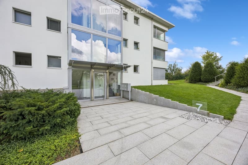 Modern two-story house with white exterior, large glass windows, and a raised ground floor entrance. The house features a well-maintained lawn, trimmed bushes, and a paved pathway leading to the entrance.