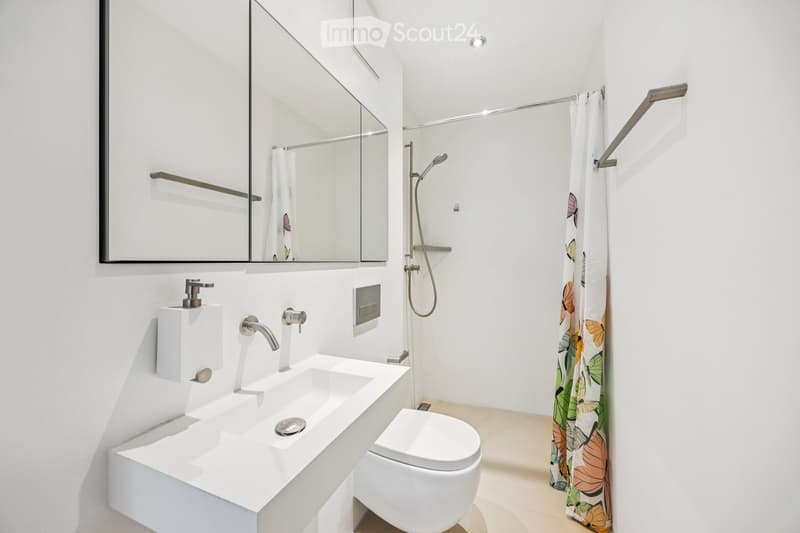Modern bathroom with white walls, a large mirror, a double sink, a toilet, and a shower with a decorative curtain.