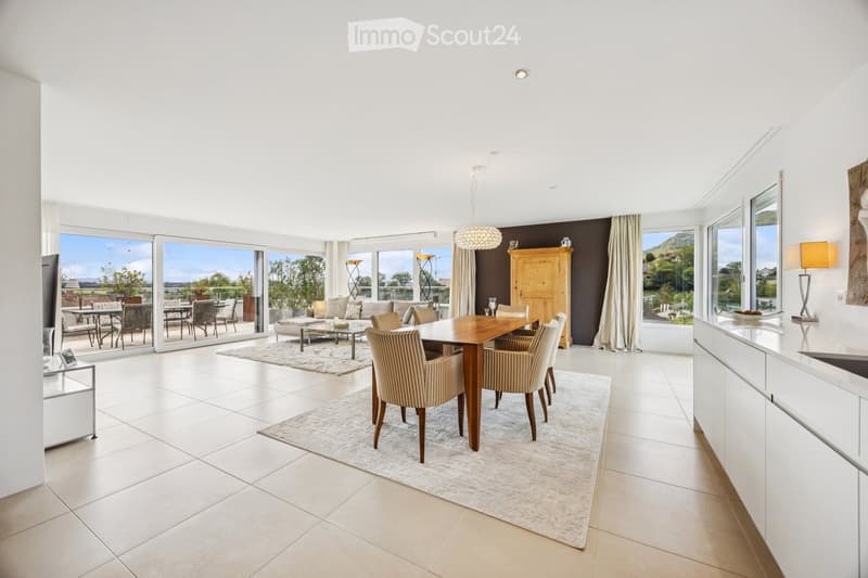 Bright, open-plan living and dining area with large windows offering scenic views, featuring a sofa, coffee table, dining table with chairs, a wooden cabinet, and lamps.