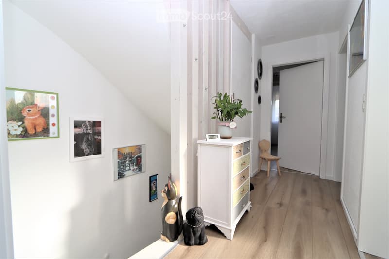 Bright hallway with white walls, wooden flooring, and several framed pictures and decorative items, including a small cabinet with drawers, a potted plant, and animal figurines.