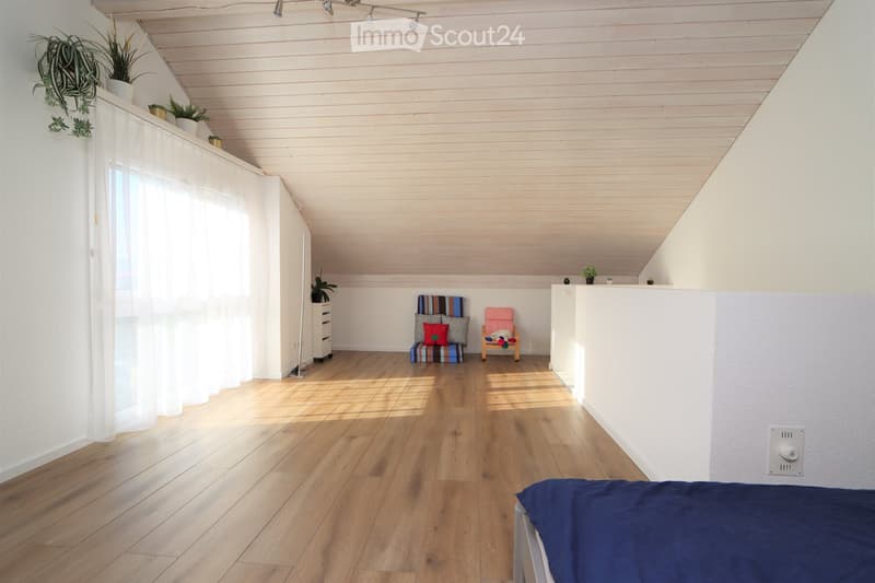 Bright, airy bedroom with wooden flooring, slanted wooden ceiling, and large window with white curtain. Features a bed with blue bedding, a small stool with colorful cushions, a wooden chair, and a white drawer unit.