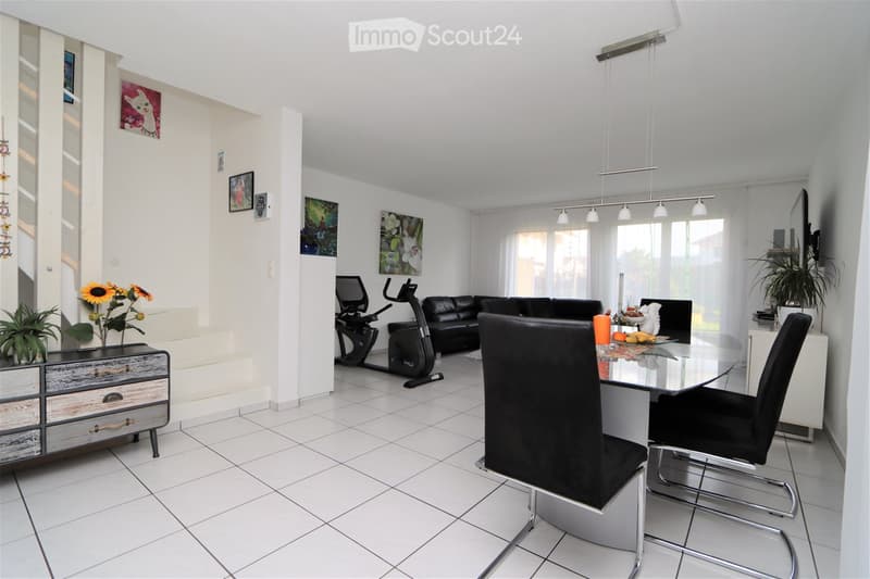 Bright, open living and dining area with white walls, tiled floor, and large windows. Features a black leather sofa, glass dining table with black chairs, exercise bike, wooden cabinet, and decorative plants.
