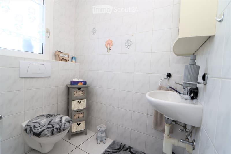A small bathroom with white tiled walls and floor, featuring a toilet with a patterned seat cover, a sink with a modern faucet, a small cabinet with woven baskets, and a decorative toy on the floor.