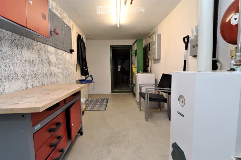 A well-lit utility or workshop room with a wooden workbench, red cabinets, a black chair, a refrigerator, and a shovel leaning against the wall.
