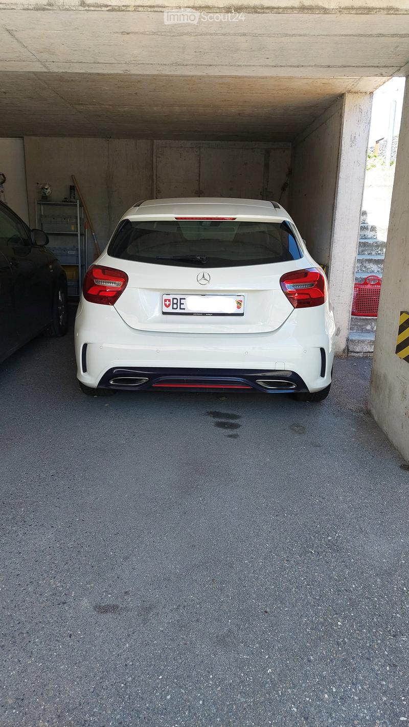 Underground garage with a white Mercedes-Benz car parked inside, concrete walls and ceiling, and a small shelf with items on the left side.