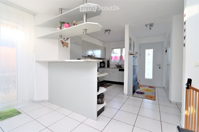 Modern kitchen with white cabinets, granite countertops, and a central island with shelves. Includes a refrigerator, microwave, sink, and decorative items on shelves.