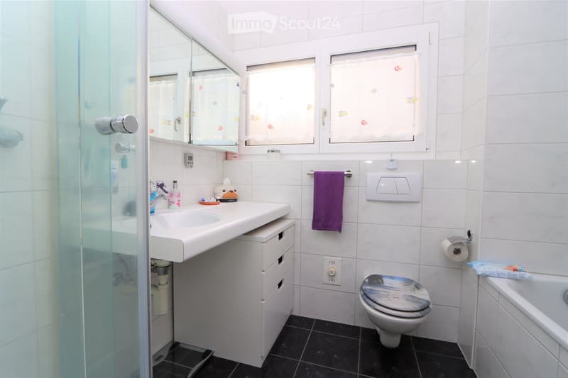 Modern bathroom with white tiles, black floor, sink with mirror, toilet, bathtub, and a purple towel hanging on a rack.