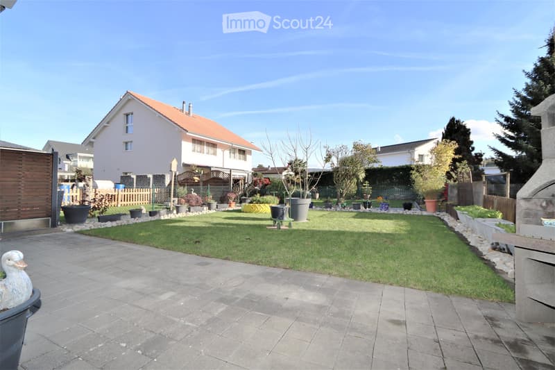 Well-maintained garden with a lawn, potted plants, a paved pathway, and a decorative duck statue. The background shows a residential house with a red-tiled roof and surrounding greenery.