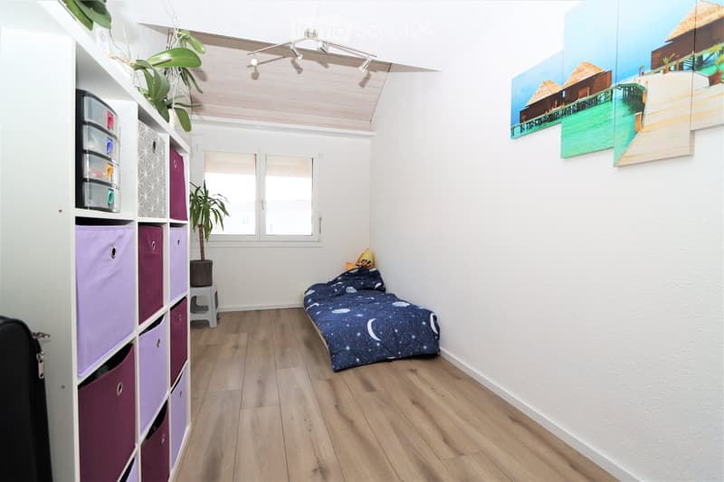 Cozy bedroom with wooden flooring, a bed with a galaxy-patterned duvet, a storage unit with colorful bins, a small stool, and a potted plant near the window.
