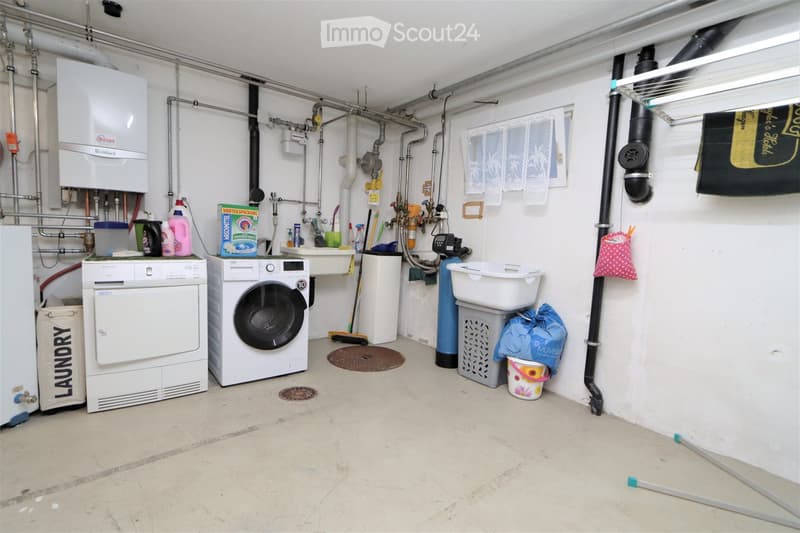 Utility room with washing machine, dryer, water heater, and various cleaning supplies. Laundry basket and bags are visible, along with a laundry sign on the wall.