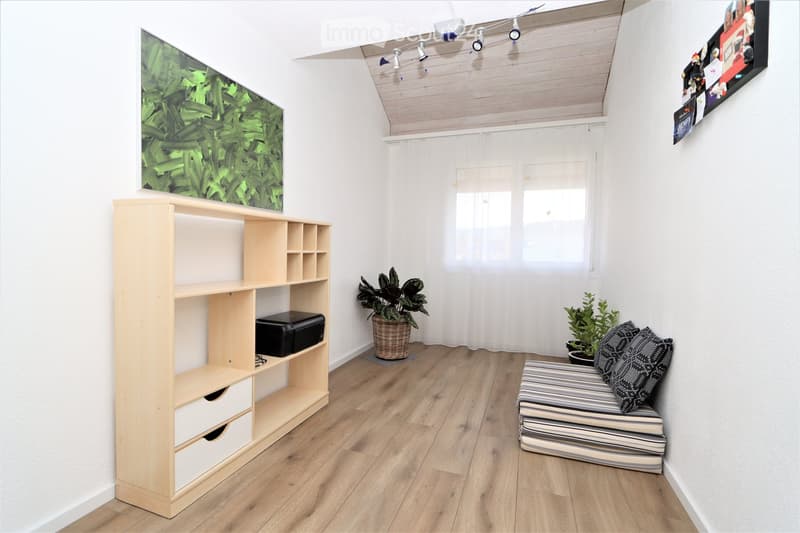 A modern, well-lit room with wooden flooring, white walls, and a slanted ceiling. Features include a wooden shelf with compartments, a small stool with patterned cushions, potted plants, and artwork on the walls.