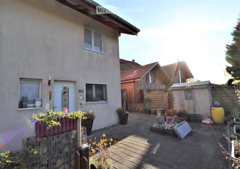 Garden area with paved patio, potted plants, wooden fence, and small shed. House entrance with decorative window boxes and owl figurines.