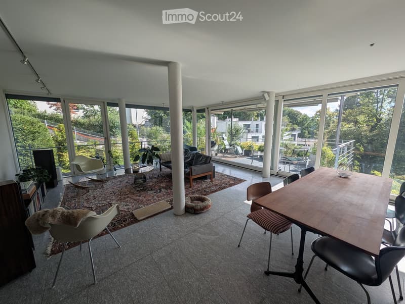 Bright living and dining area with large windows and a balcony, featuring a sofa, dining table, chairs, and plants.