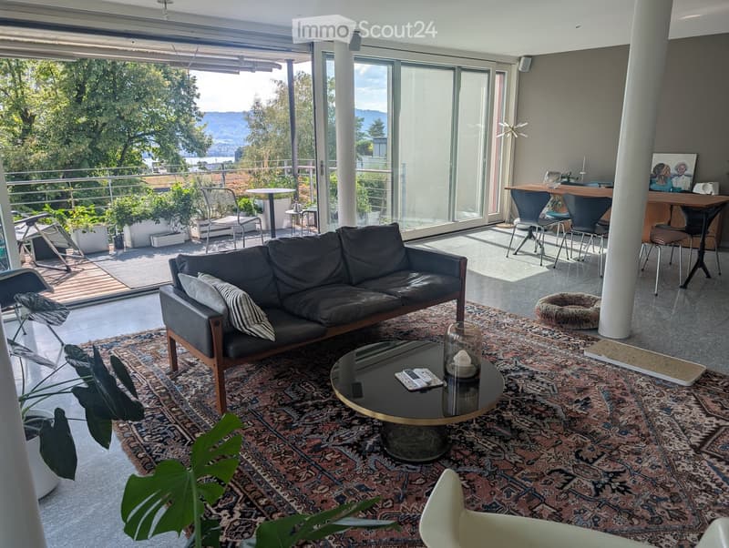 Modern living room with large windows offering a scenic view, featuring a dark leather sofa, coffee table, chairs, and a small table with stools in the adjacent area.
