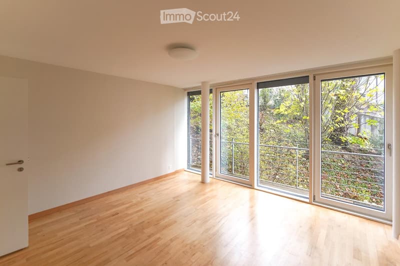 Empty room with light wooden flooring, white walls, and large sliding glass doors leading to a balcony with a view of greenery.