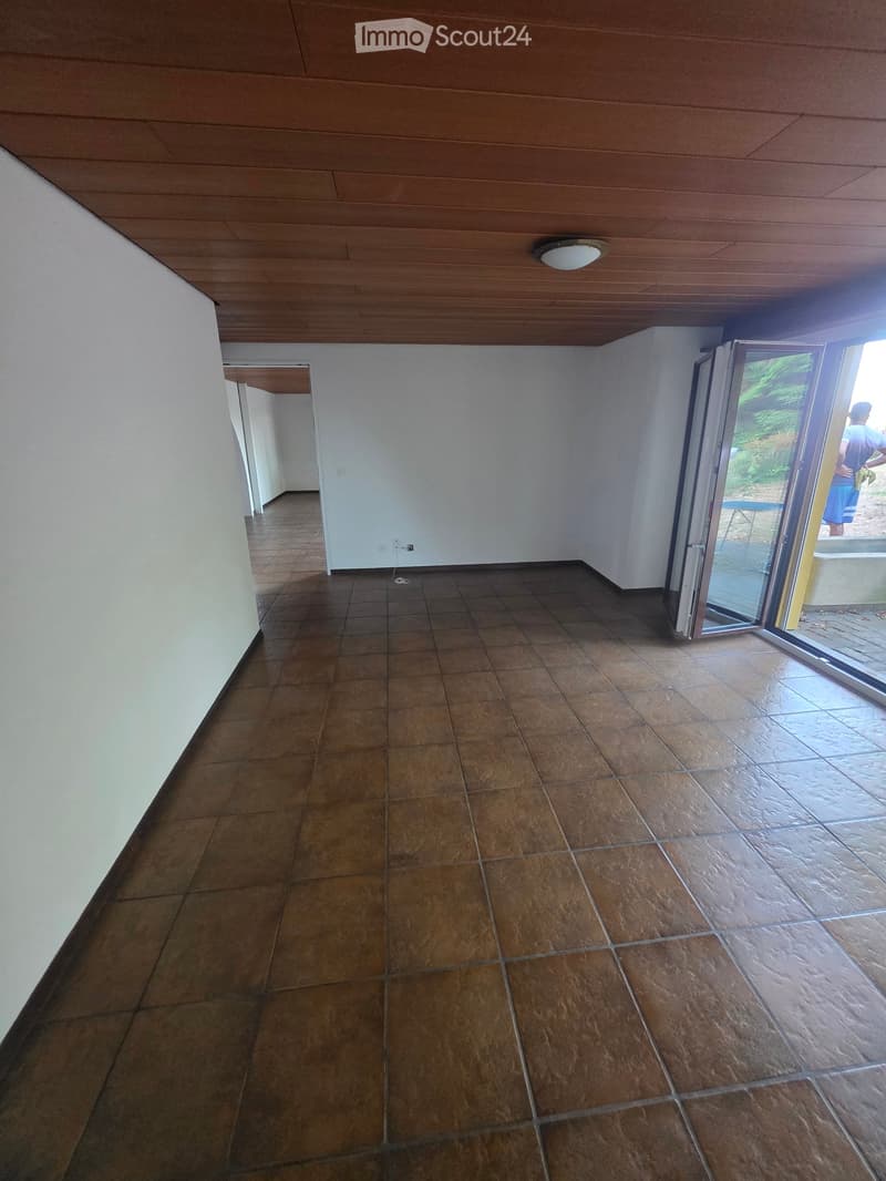 Empty room with tiled floor, white walls, wooden ceiling, and large sliding glass door leading outside.