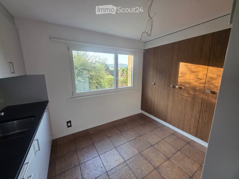 Empty kitchen with wooden cabinets, tiled floor, and large window overlooking greenery.