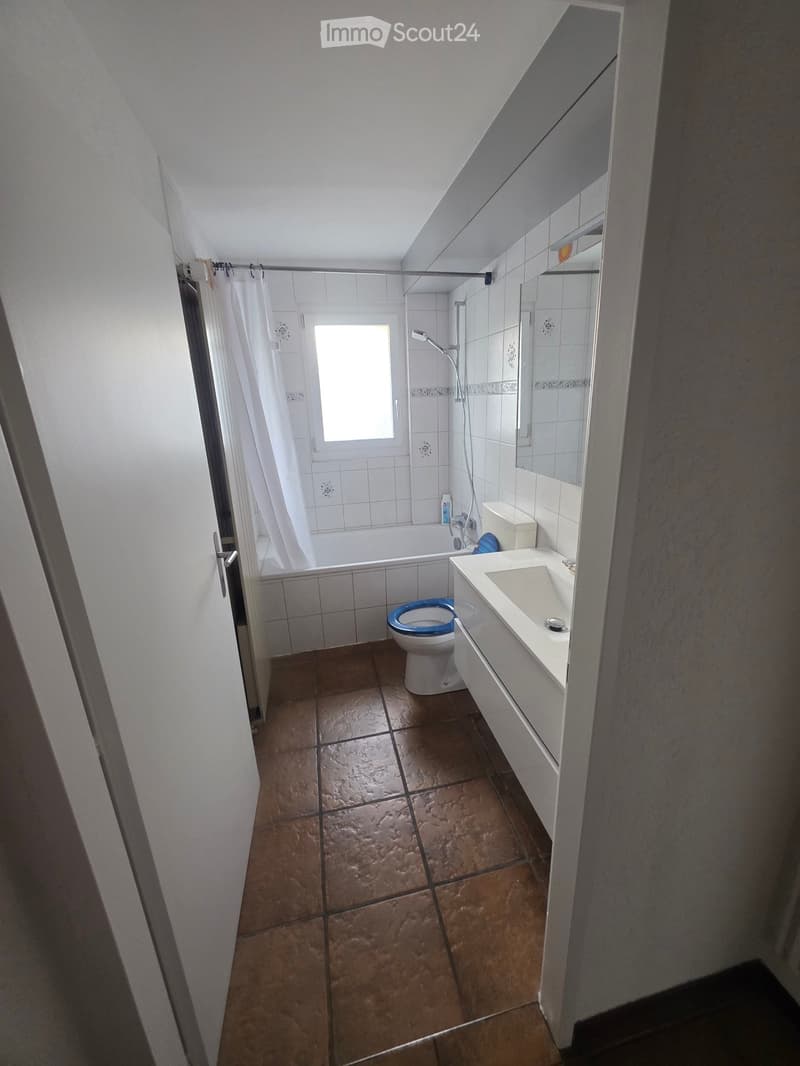 Bathroom with brown tiled floor, white tiled walls, toilet with blue seat, sink, bathtub, and shower area with white curtain.