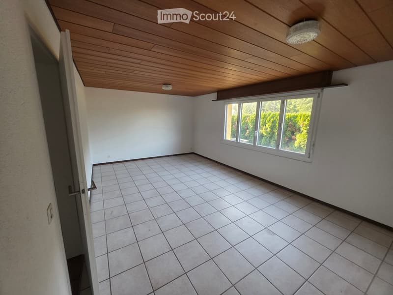 Empty room with white tiled floor, white walls, wooden ceiling, and large window with view of greenery.