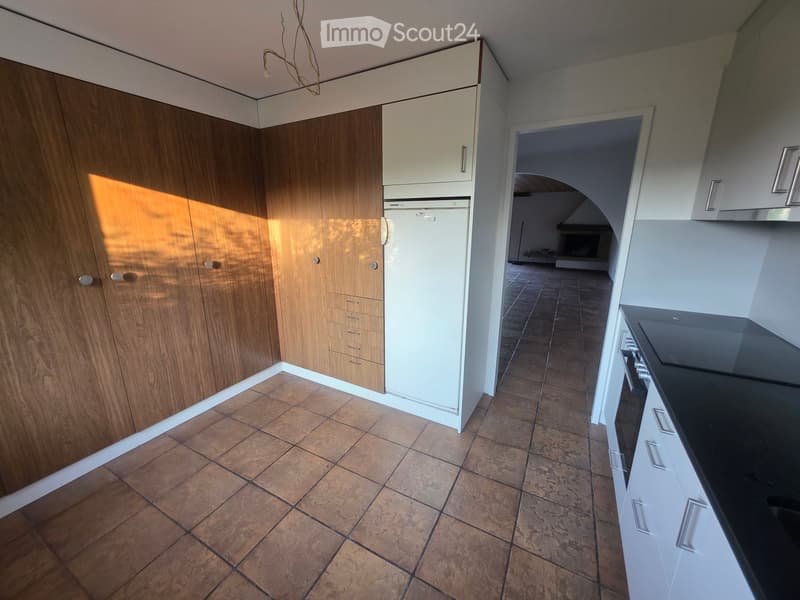 Modern kitchen with wooden cabinets, white refrigerator, black countertop, and tiled floor. An open doorway leads to a living area with a fireplace.