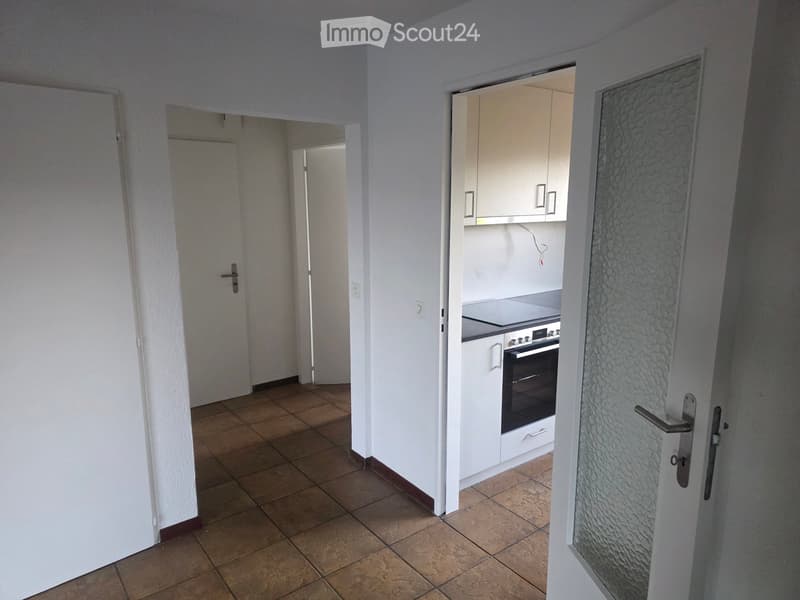 Empty hallway with tiled floor, white walls, and doors leading to other rooms, including a kitchen area with cabinets and an oven.