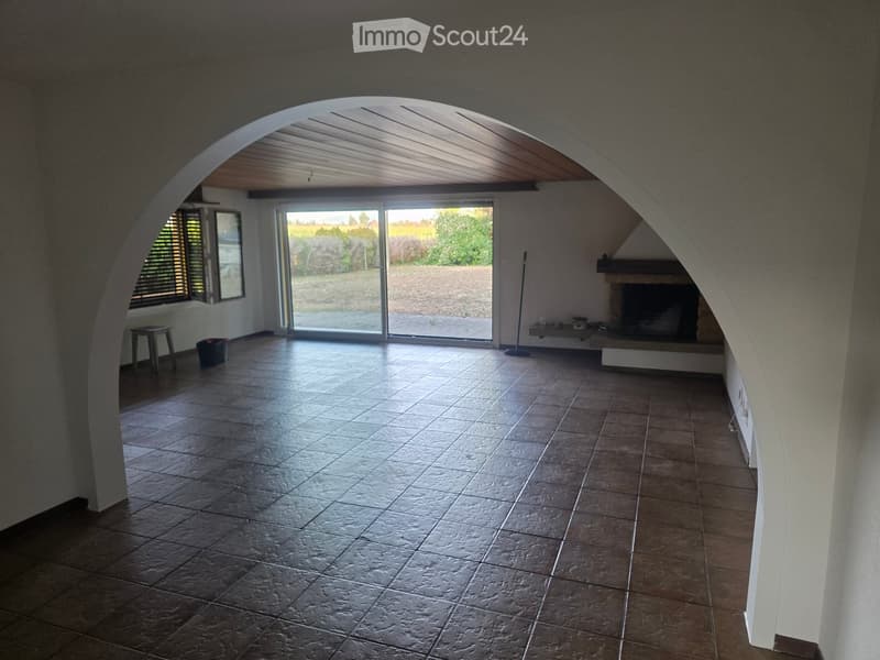 Empty room with tiled floor, arched ceiling, large sliding glass door leading to outdoor area, fireplace, small table, and broom.
