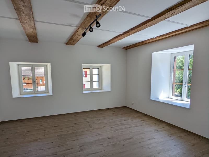 Empty room with wooden flooring, white walls, and three windows, including a large window with a view of greenery.