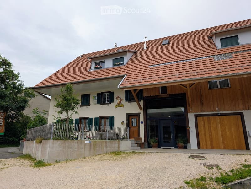Two-story building with red-tiled roof, white walls, and wooden garage entrance. Features multiple windows with shutters, a small garden area, and a gravel driveway.