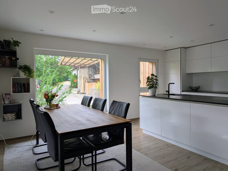 Modern open-plan dining and kitchen area with a wooden dining table, black chairs, white kitchen cabinets, and a sink.