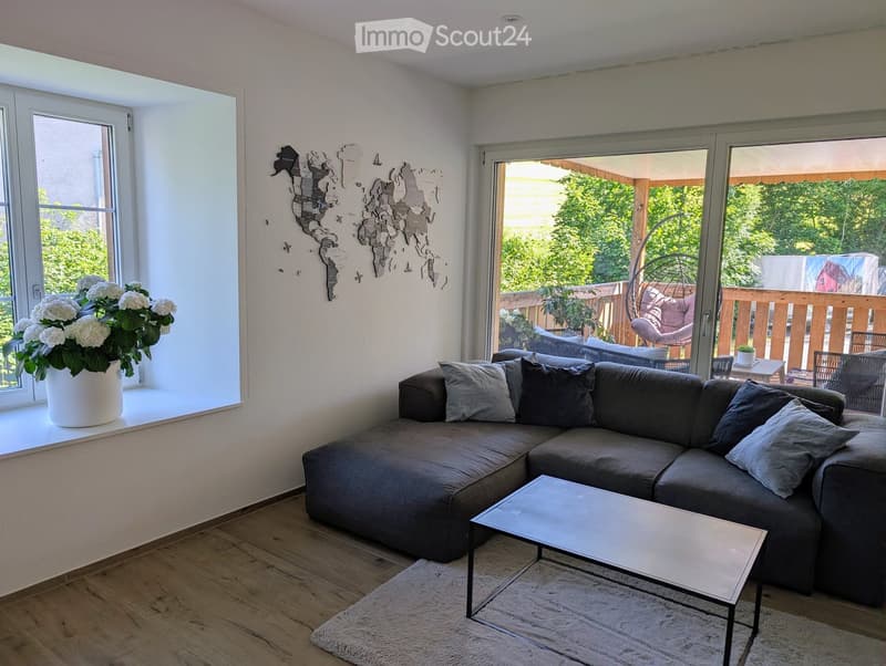 Modern living room with a grey sectional sofa, coffee table, and a large window with a flower pot on the sill. The room has wooden flooring and a view of a balcony with outdoor furniture.