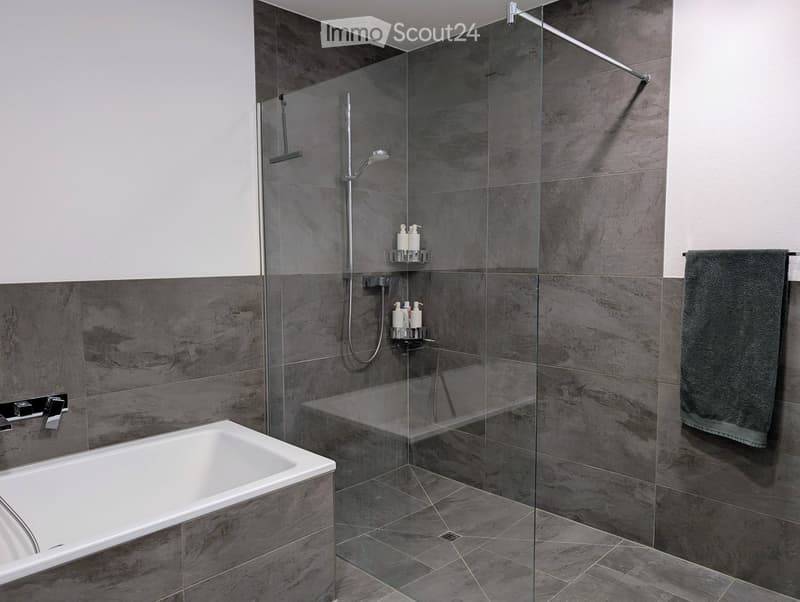 Modern bathroom with a white bathtub, glass-enclosed shower, dark grey tiles, and a towel on the side.