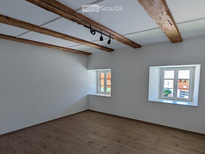 Empty room with wooden flooring, white walls, exposed wooden beams on the ceiling, and two windows with white frames.