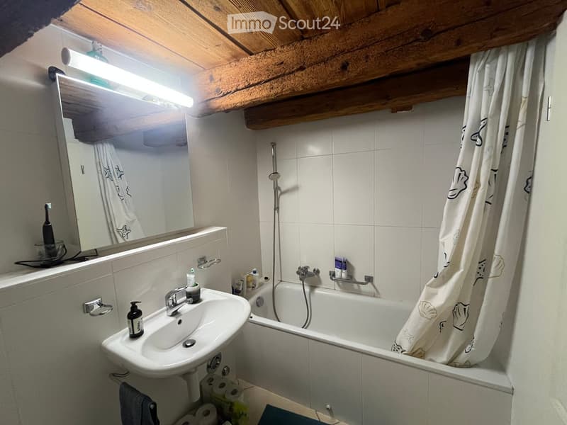 Bathroom with a sink, bathtub, mirror, and shower curtain. Exposed wooden beams on the ceiling and a shelf with toiletries.