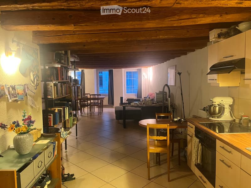Open-plan living and dining area with exposed wooden beams, tiled floor, and modern kitchen. Features a sofa, dining table, bookshelf, and kitchen appliances.