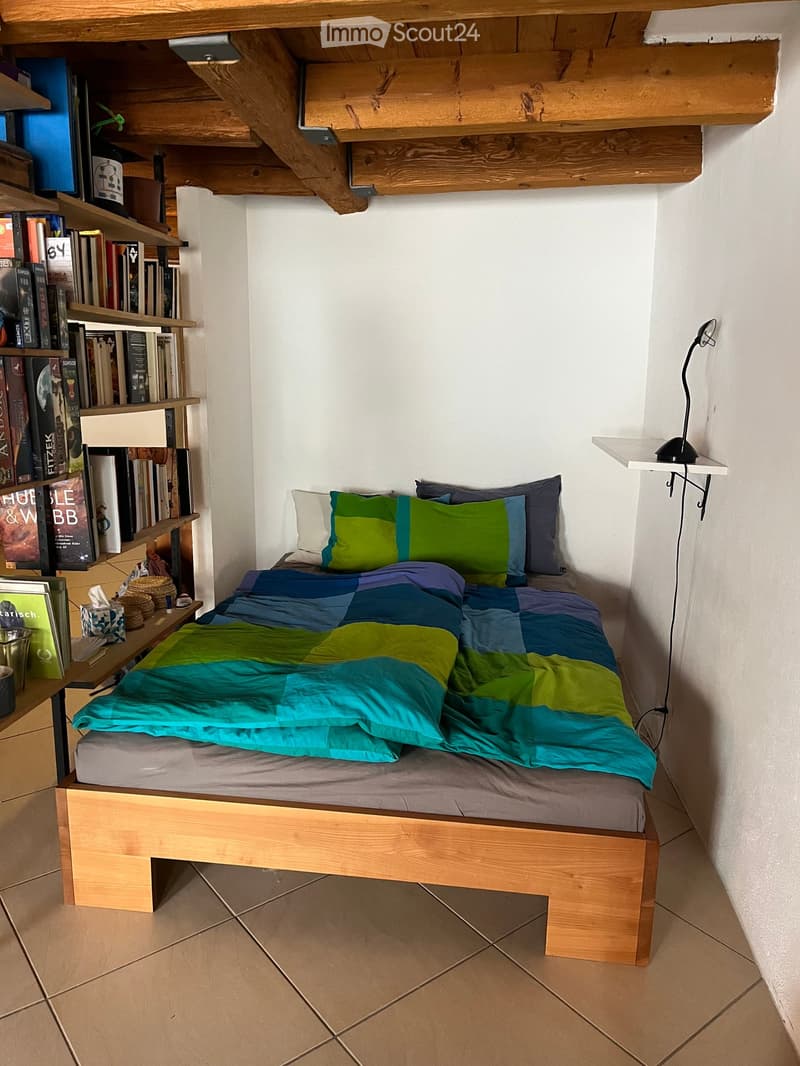 A small bedroom with a wooden bed, colorful bedding, a bookshelf filled with books, and a small table with a lamp.