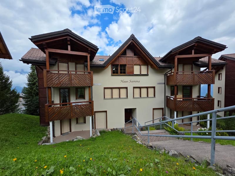Three-story apartment building, beige walls, wooden balconies, name sign 'Haus Samina', staircase leading to entrance, green surroundings