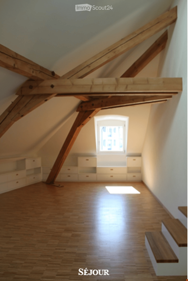An attic room with exposed wooden beams, wooden flooring, built-in white cabinets, and a window letting in natural light.