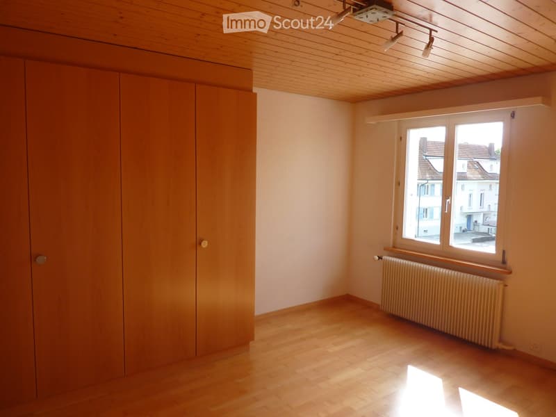 Empty room, wooden floor, large wooden wardrobe, white walls, wooden ceiling, window with white frame, radiator beneath the window