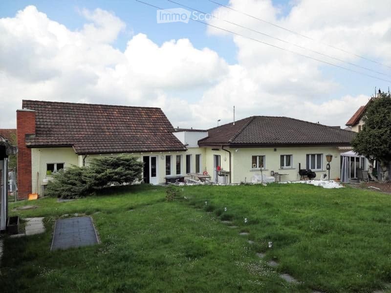 Two-story house with beige walls, red-tiled roof, and multiple windows. The front yard is grassy with a small tree and outdoor furniture.