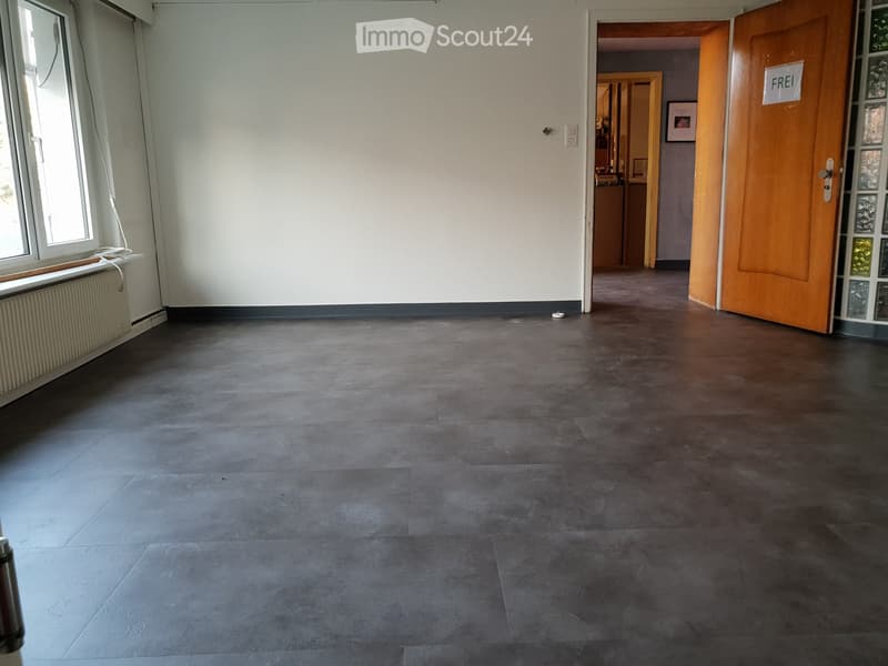 Empty room with dark grey tiled floor, white walls, and a large window with a radiator below it. An open wooden door leads to another room.