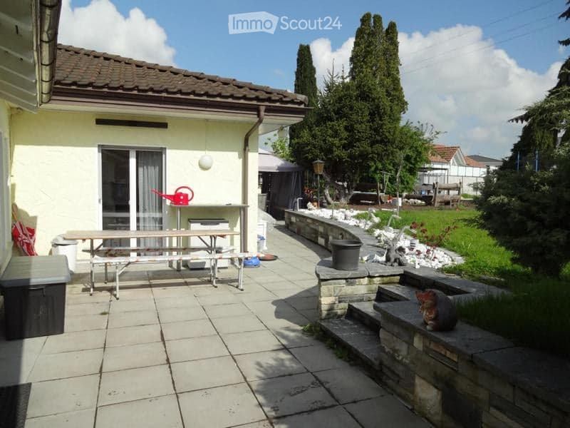Outdoor patio area with tiled flooring, a wooden picnic table, a stone staircase leading to a garden, and surrounding greenery.