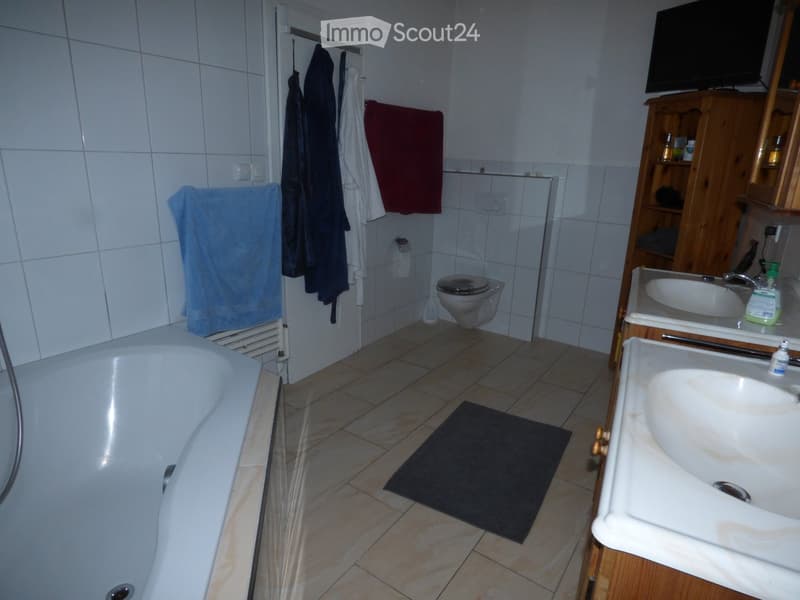 Bathroom with a bathtub, toilet, sink, mirror, and wooden cabinet. Towels are hanging on the wall, and a black bath mat is on the floor.