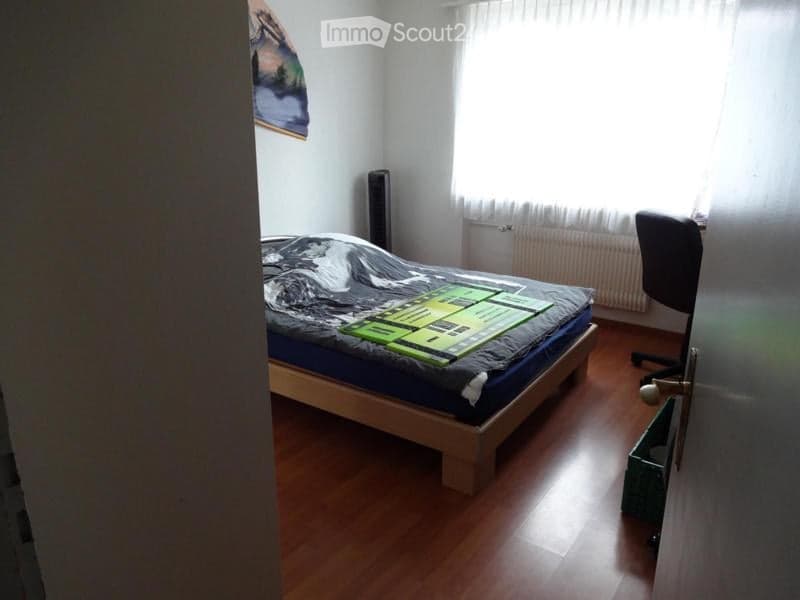 Bedroom with wooden floor, bed with green and black duvet, chair, radiator, speaker, and a painting on the wall.
