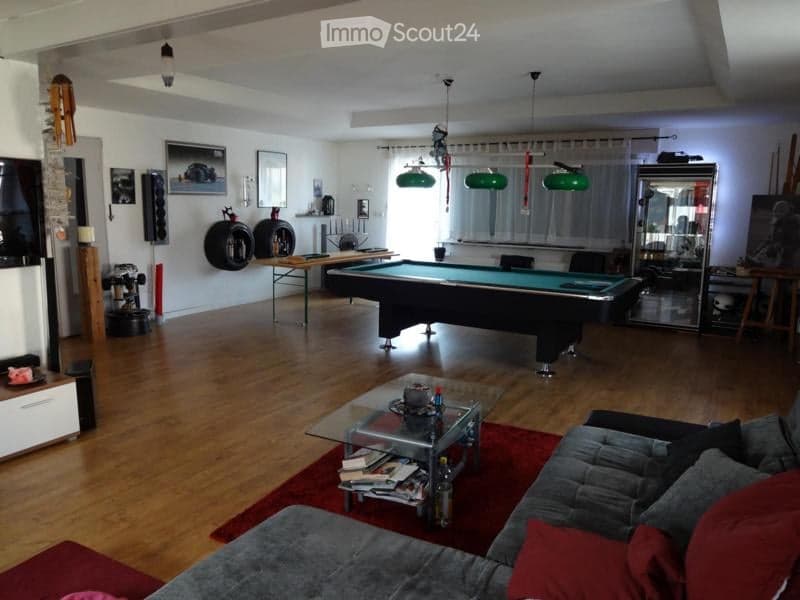 Living room with wooden flooring, a grey sofa, a glass coffee table, a pool table, and a large mirror. The room features several hanging green lamps and a variety of decorative items on shelves and walls.
