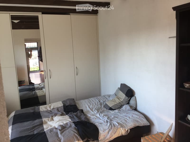 A small bedroom with a neatly made bed, a large white wardrobe, and a dark wooden shelf. The bed features a patterned duvet and pillows, and the room has exposed wooden beams on the ceiling.