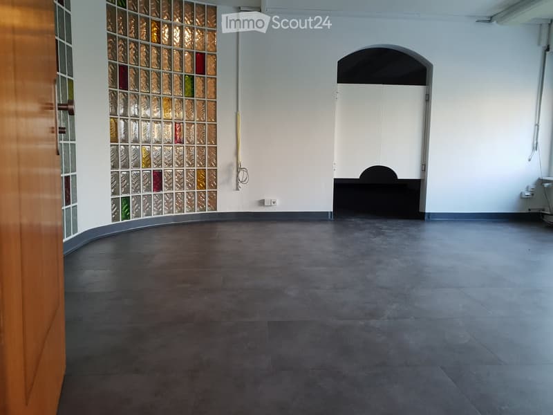 Empty room with dark grey tiled floor, white walls, and a partially visible wooden door with decorative glass panels. There is an arched doorway leading to another space.