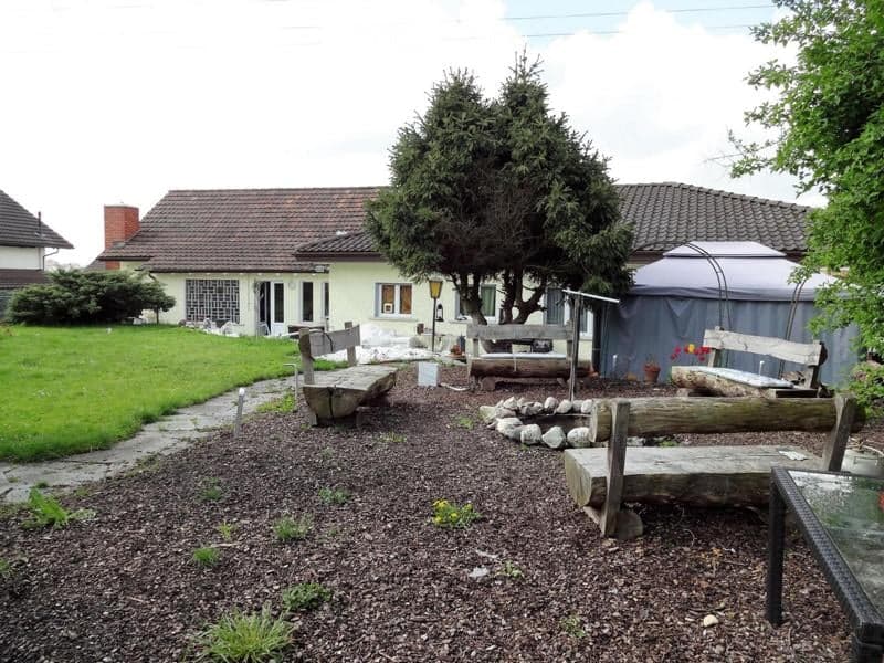 Garden area with benches, a fire pit, and a small table, surrounded by a house with a tiled roof and large windows.